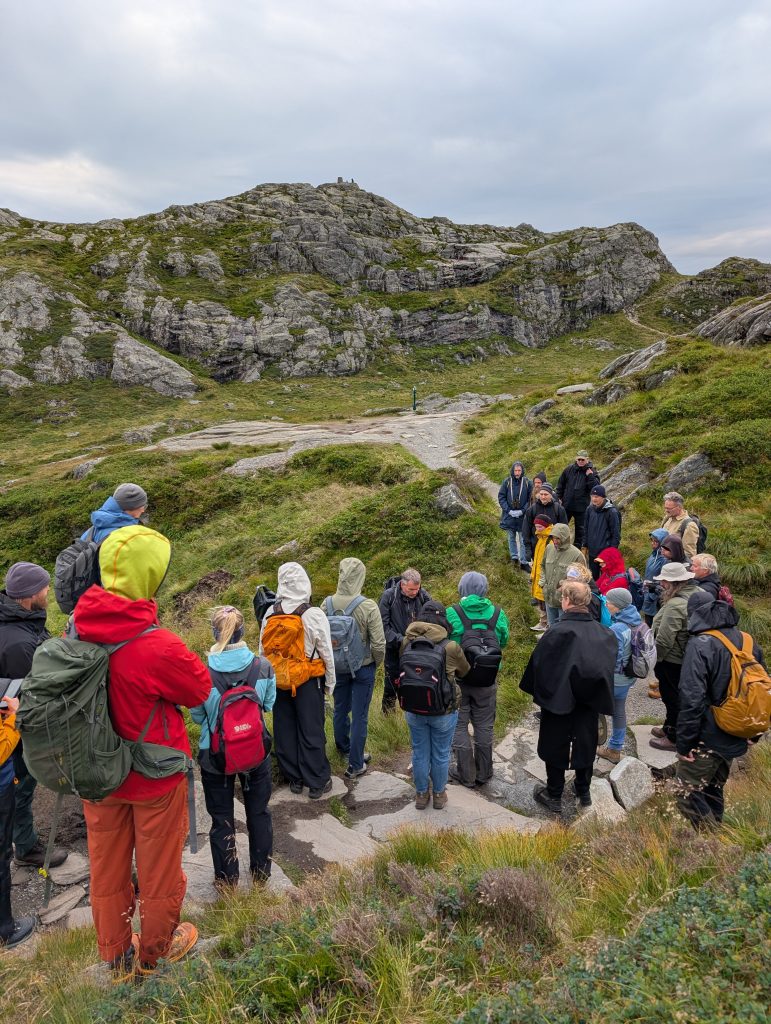 A group of people in raincloths, with their backs to the camera, standing in mountains with green grass under a cloudy sky.