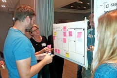 Three people standing next to a board with pink post-it notes on it. Photo.