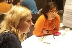 Two women sitting at a table leaning forward. Photo.