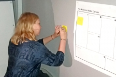 Woman standing next to white board writing on a post-it. Photo.