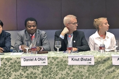 Six people in a panel, one woman holding a microphone. Photo.